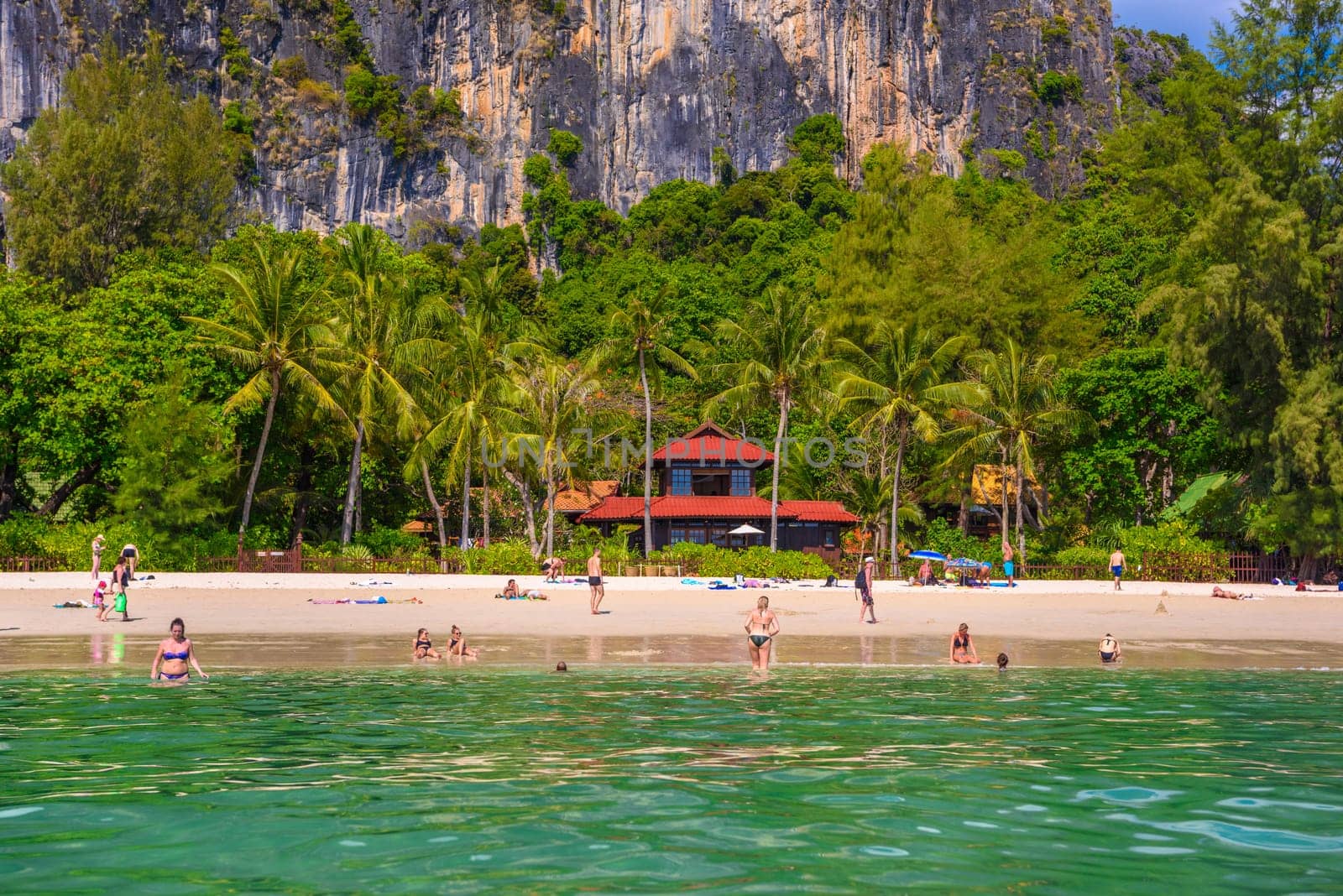 Bungalow house with red roof among coconut palms near the cliffs with people sunbathing and swimming in emerald water on Railay beach west, Ao Nang, Krabi, Thailand by Eagle2308