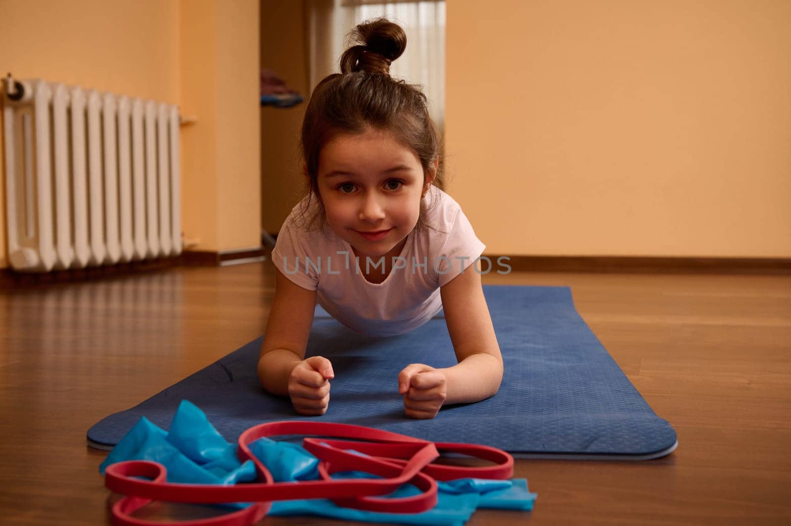 Front shot of a 5-6 years old Caucasian determined sporty little child girl, looking at camera, doing plank exercise on a blue fitness mat while working out, stretching body, practicing yoga at home