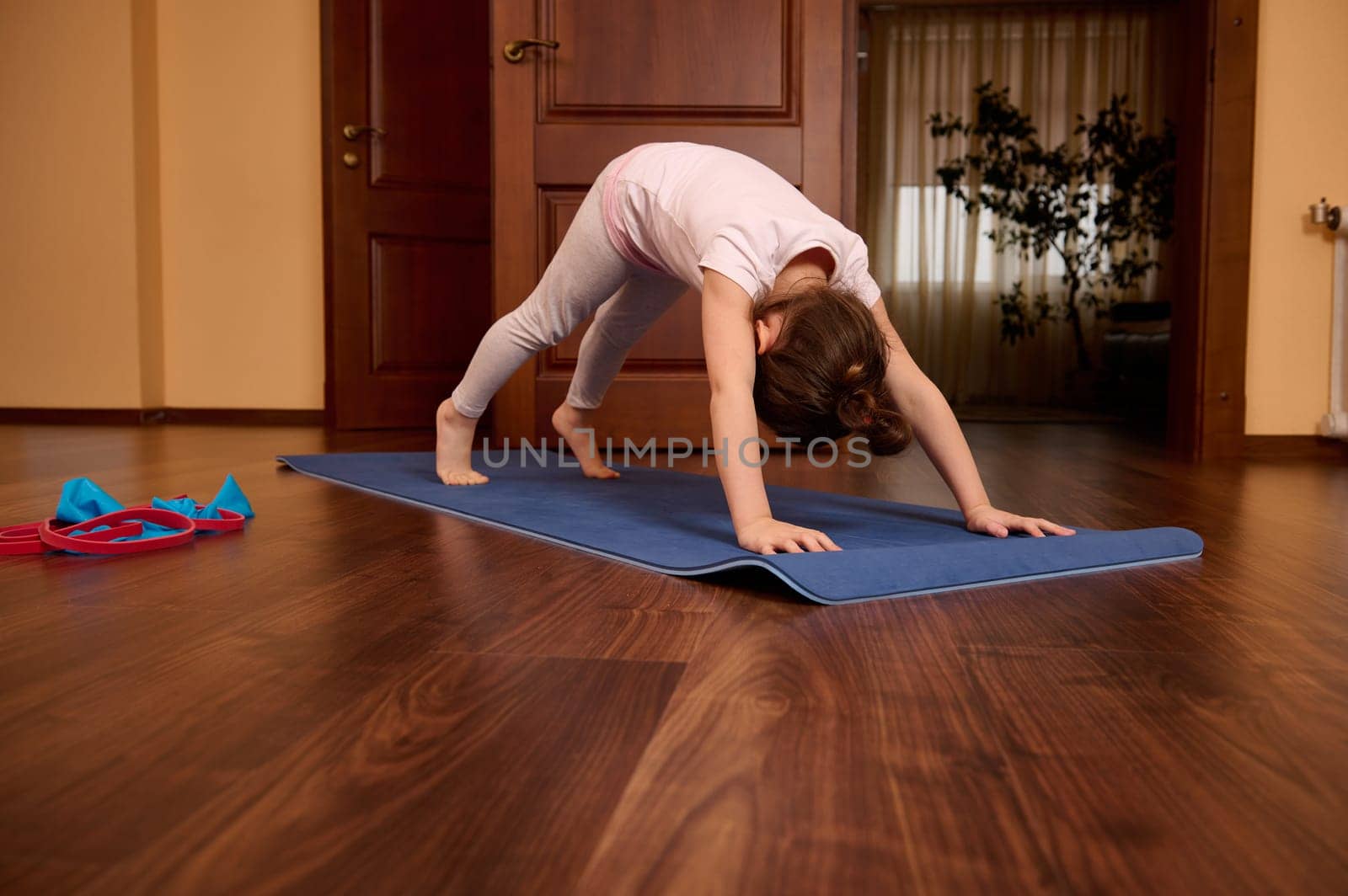 Caucasian 5-6 years old cute little girl, adorable sporty kid stretching her body in downward facing dog pose, practicing yoga in cozy home interior. Yoga practice. Sport. Health and active lidestyle