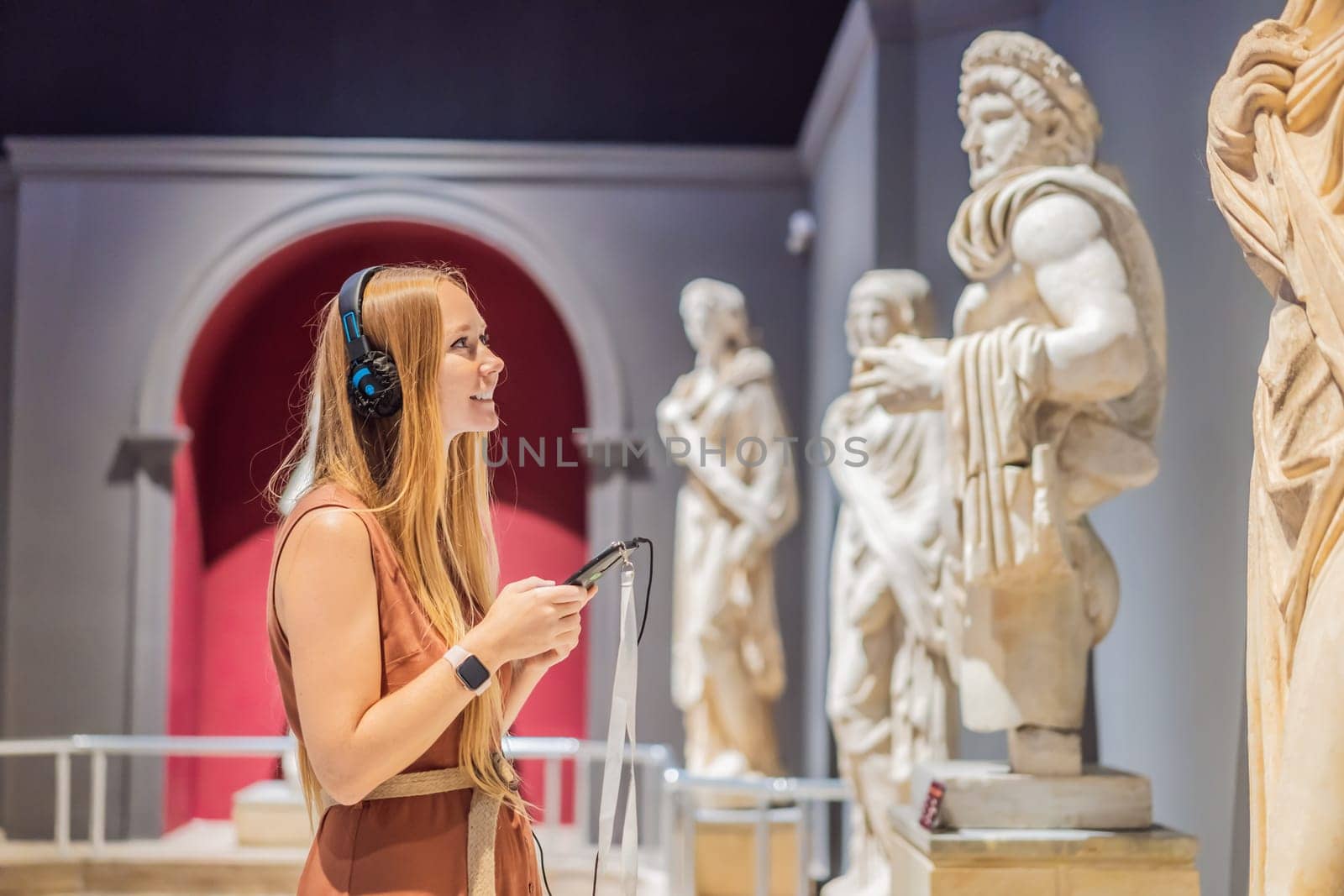 Portrait of contemporary young woman looking at sculptures and listening to audio guide at museum exhibition.
