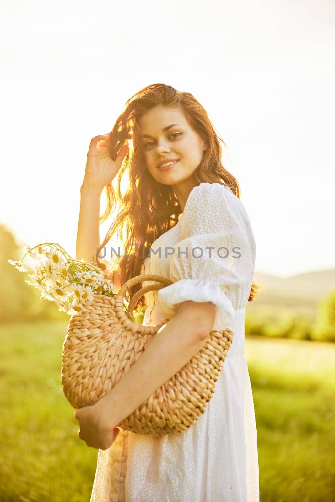 a laughing woman with a full basket of daisies stands in a light dress against the backdrop of a sunset in a field. High quality photo