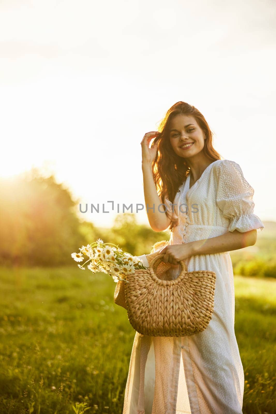 a laughing woman with a full basket of daisies stands in a light dress against the backdrop of a sunset in a field. High quality photo