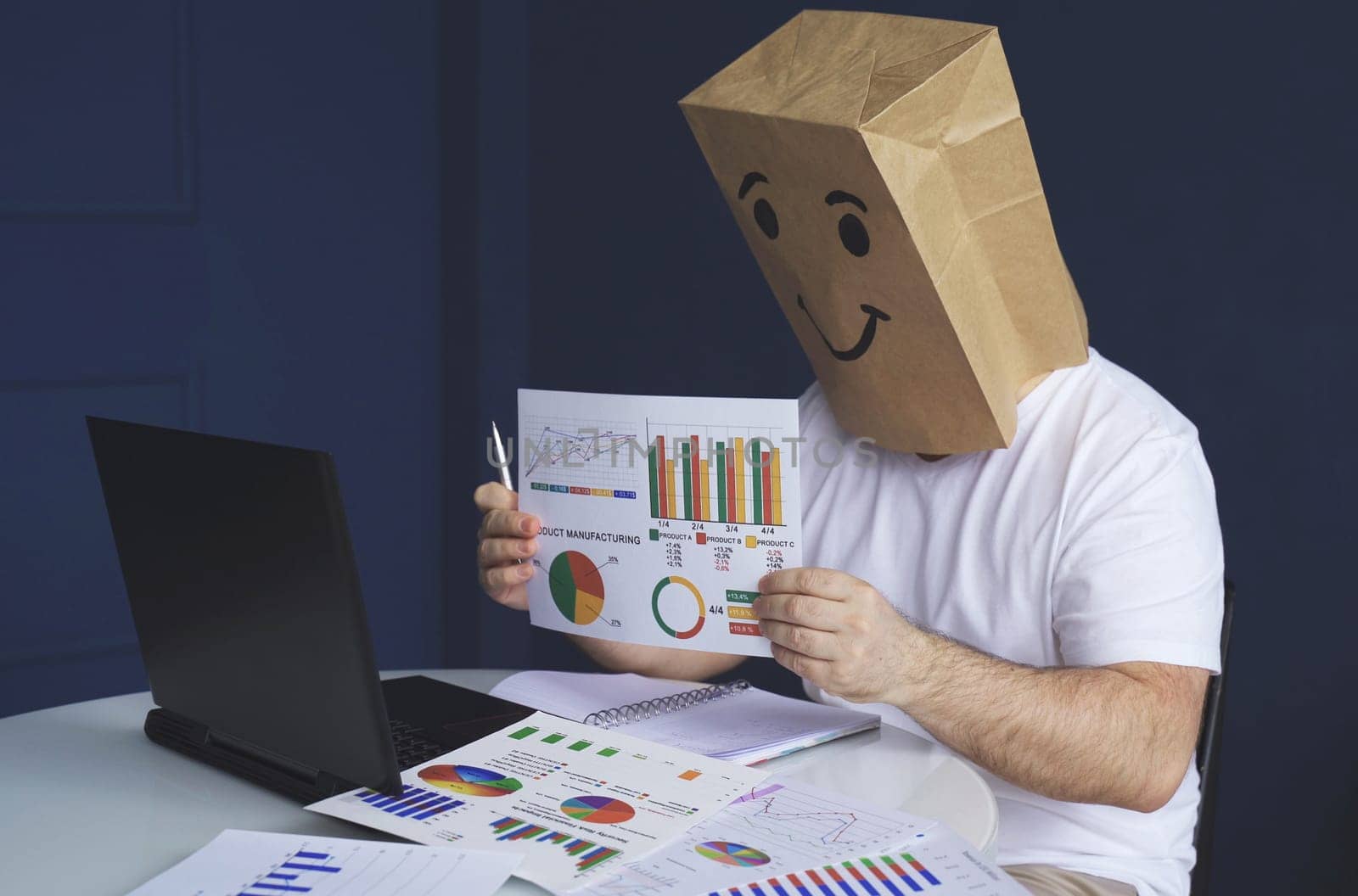 A man is a businessman in a white shirt with a paper bag on his head, with a joyful smiley face drawn, conducts a video conference or training via video link on a laptop. Emotions and gestures.