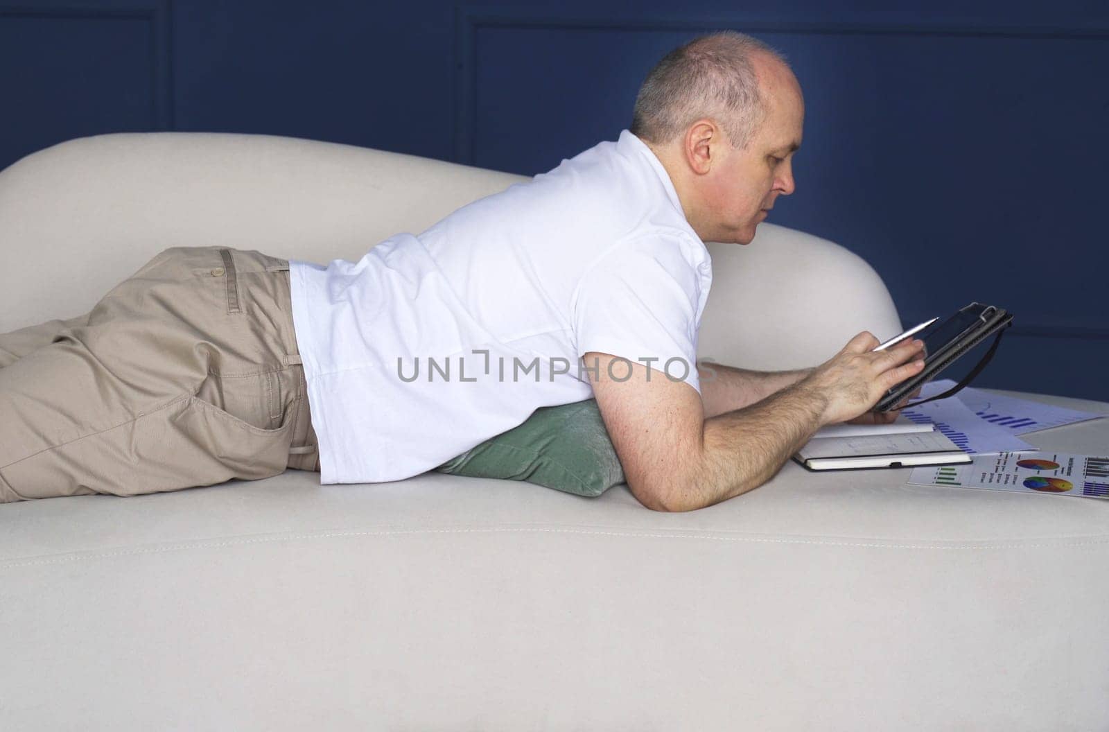 A businessman lies on a sofa and looks at data trends on charts and graphs in a tablet, makes notes in a notebook. Distant work