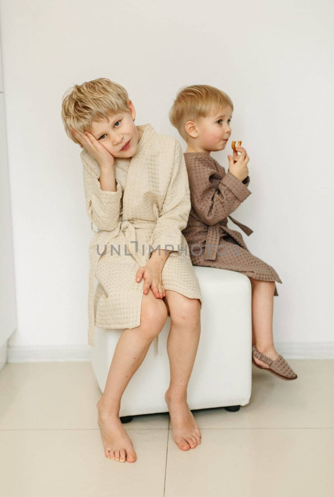 Two boys in bathrobes are sitting on a pouf in a white room.
