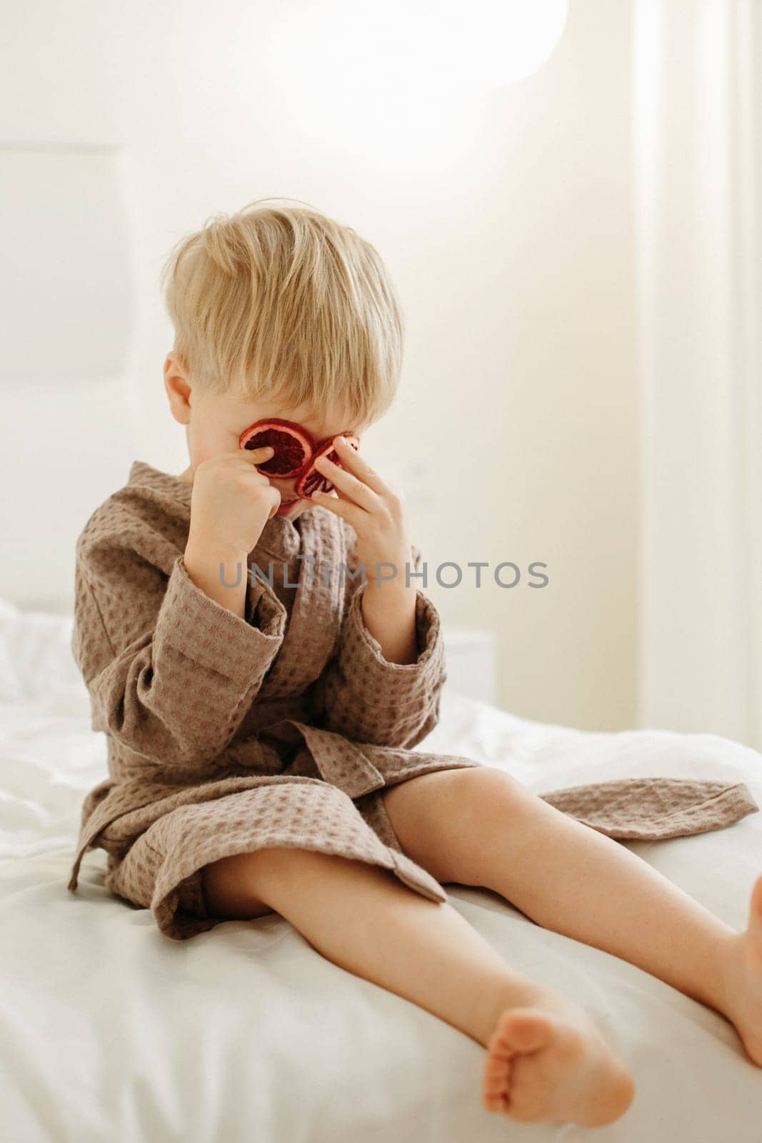 A boy in a brown bathrobe sits on the bed, plays - he closed his eyes with candied oranges. Vertical frame.