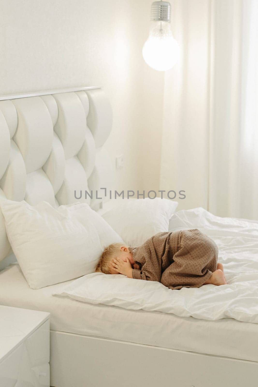 A boy in a brown bathrobe, lies on a white bed, covers his face with his hands.