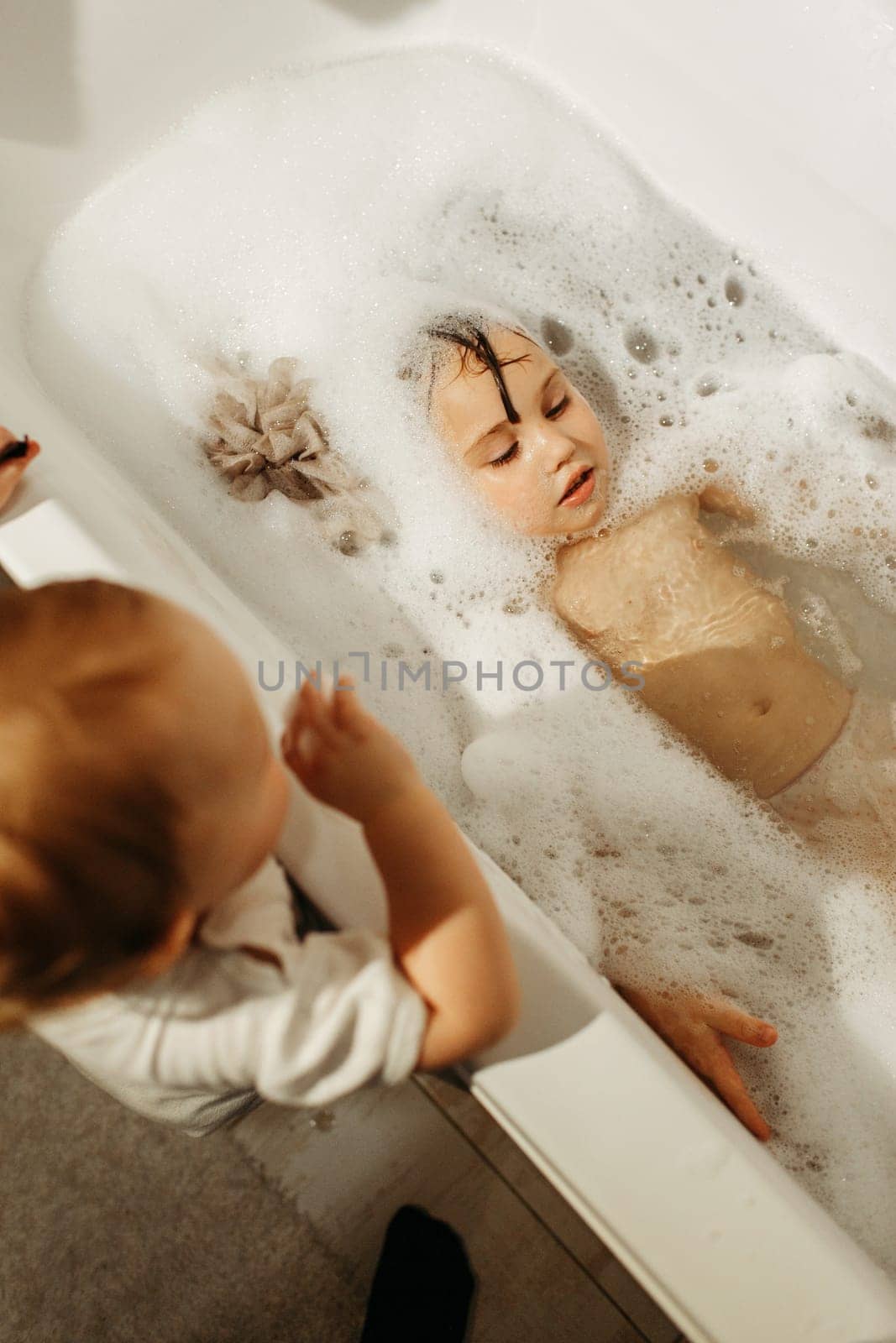 A happy girl swims in a bathtub full of foam with her little sister standing next to her. Only the face is visible from the foam.
