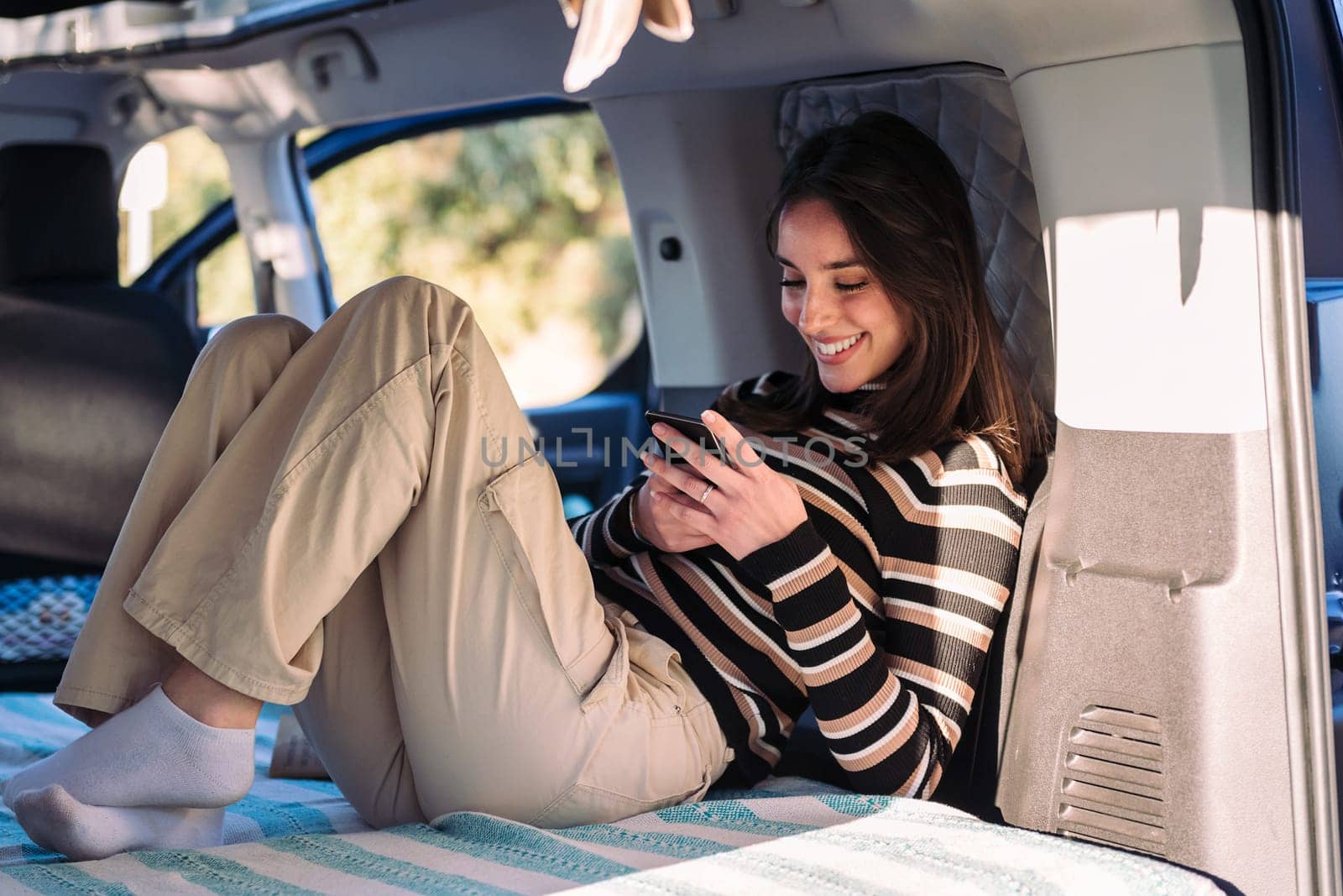 smiling young caucasian woman relaxing in camper van using a mobile phone, concept of road trip adventure and van life relaxation