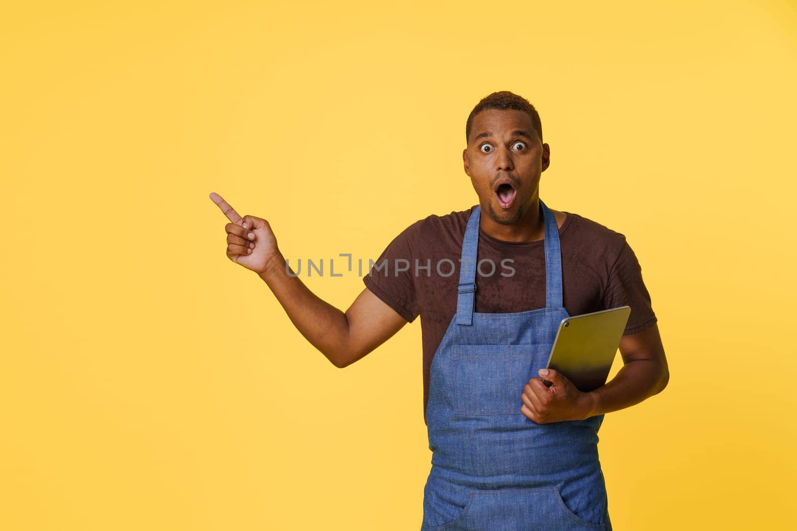 Amazed young African American cook in blue apron, searching for recipes online on tablet PC. Cook holding tablet in one hand and gesturing towards the copy space on bright yellow background with other hand. by LipikStockMedia