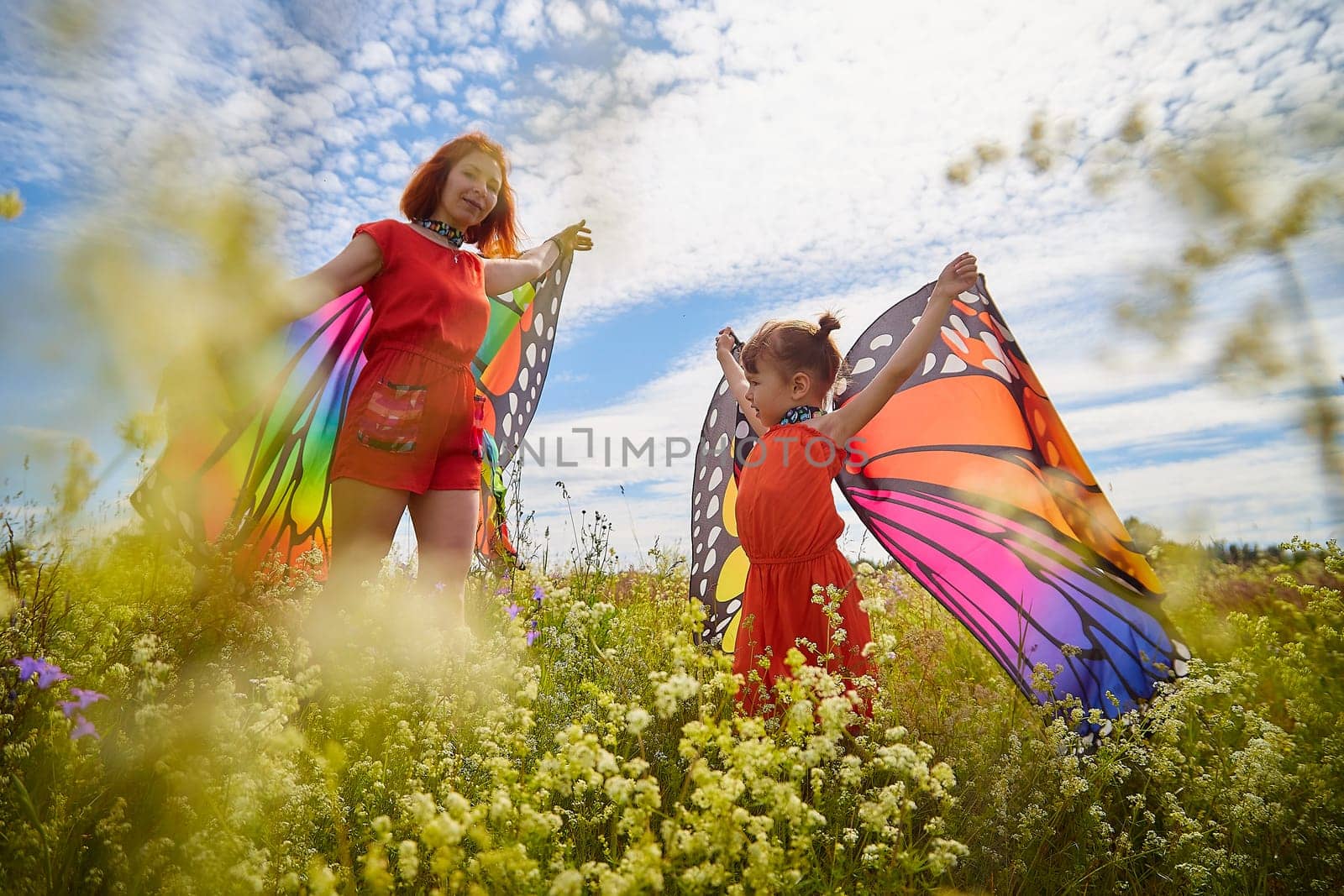 Happy female family with red haired mother and daughter with bright butterfly wings having fun on green and yellow meadow full of grass and flower in sunny summer day. Concept family love by keleny