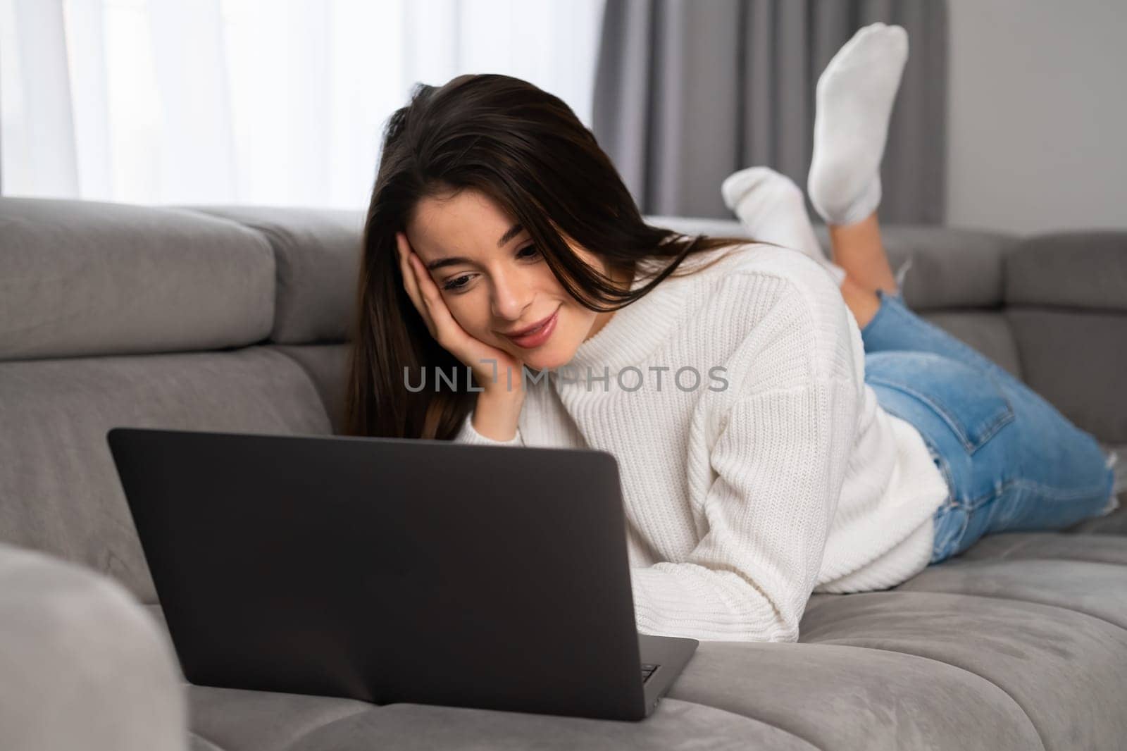 Young woman enjoys chat with friends via modern Notebook. Satisfied lady lies on couch in living room using modern laptop for online communication