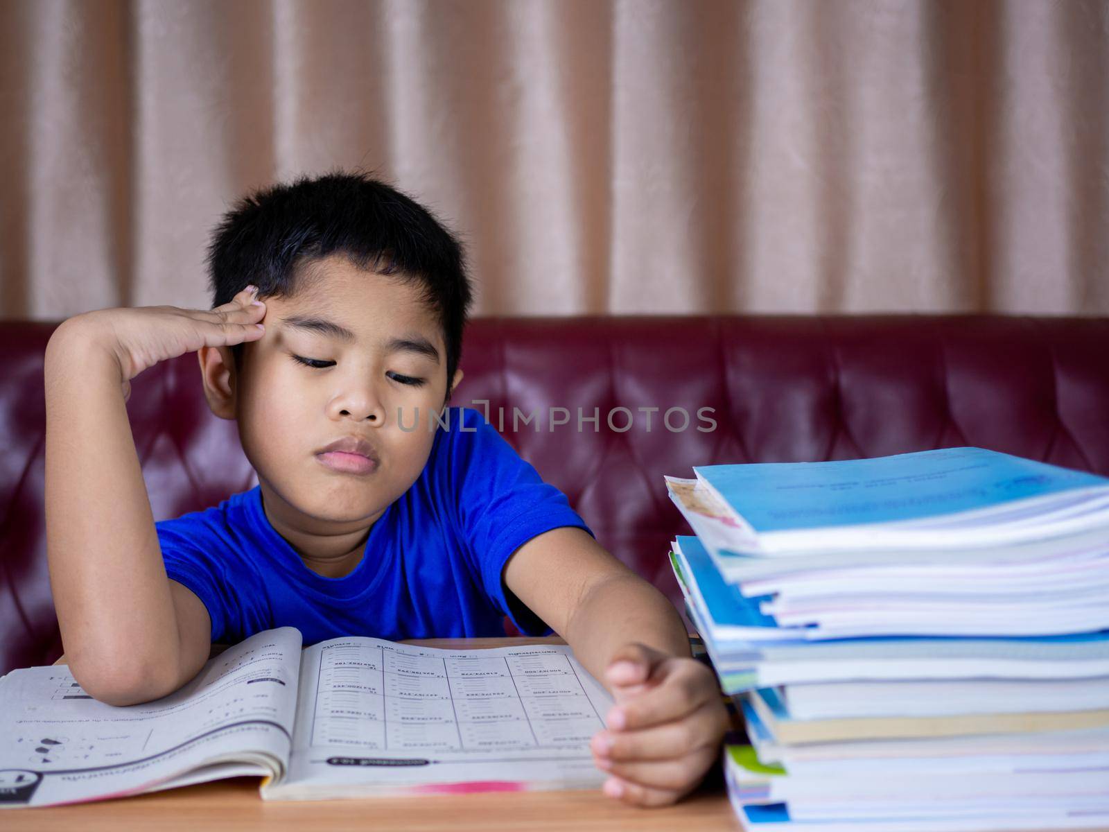 A boy is tired of reading a book on a wooden table. with a pile of books beside The background is a red sofa and cream curtains.