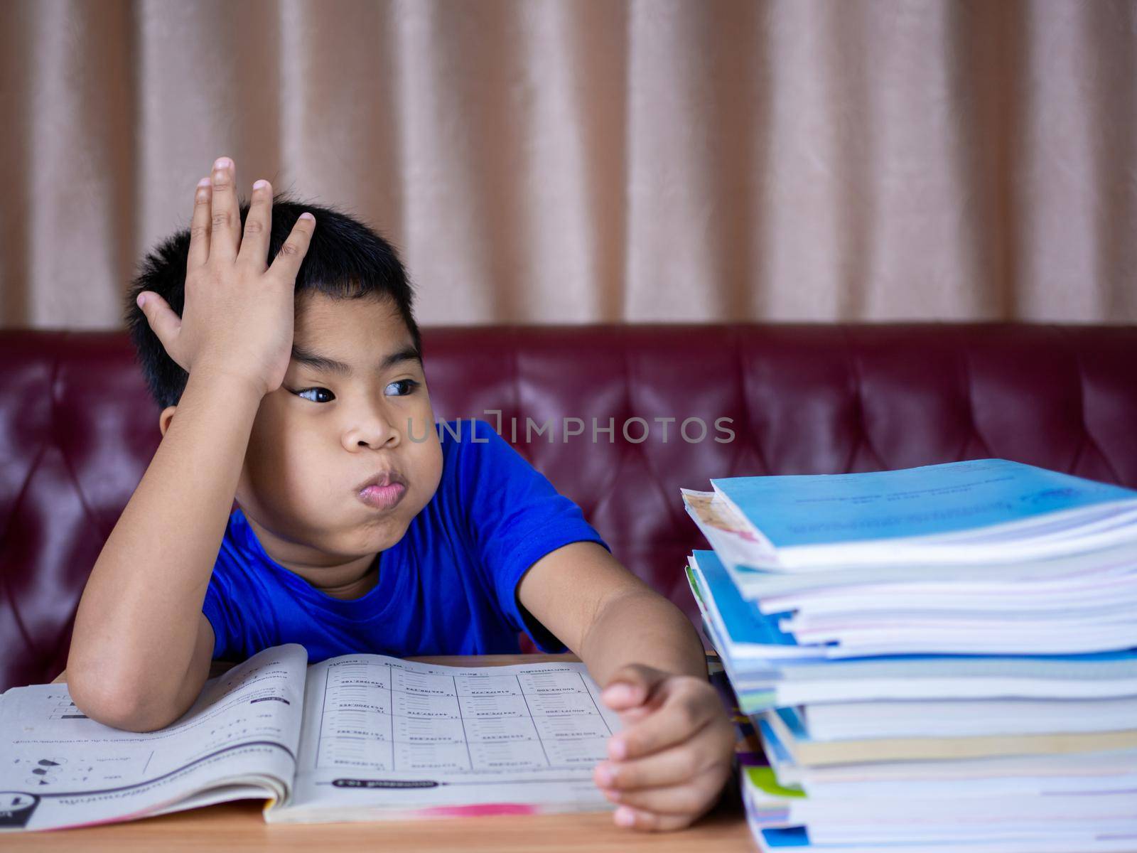 A boy is tired of reading a book on a wooden table. with a pile of books beside The background is a red sofa and cream curtains.