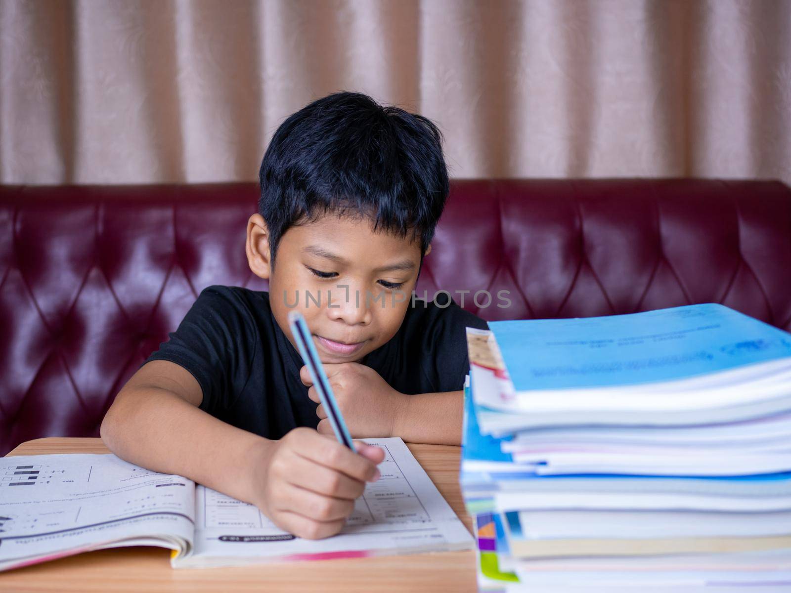 boy doing homework and reading on a wooden table with a pile of books beside The background is a red sofa and cream curtains. by Unimages2527