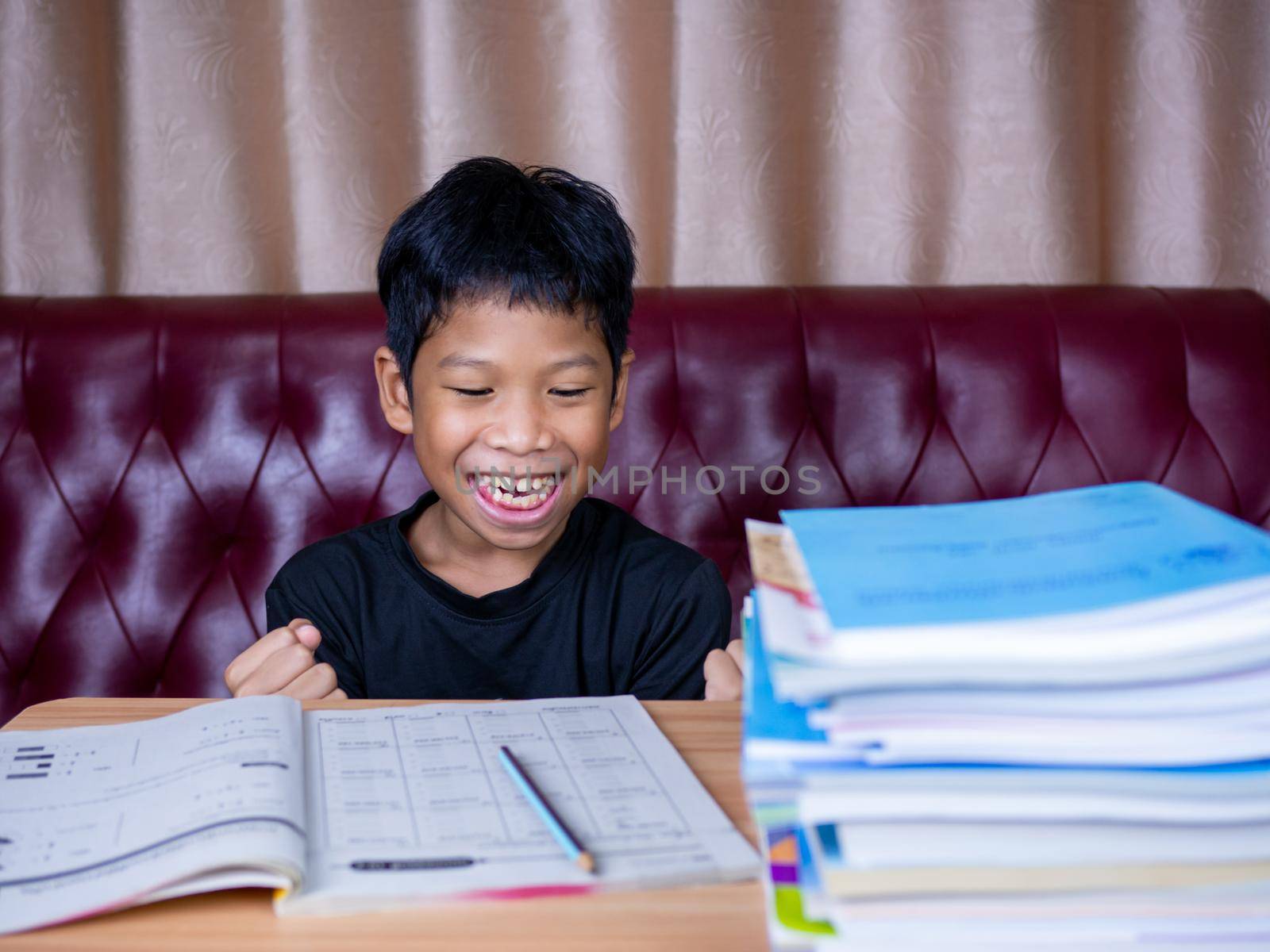 The boy was very happy to finish his homework. He sat on a wooden table and had a stack of books next to it. The background is a red sofa and cream curtains.