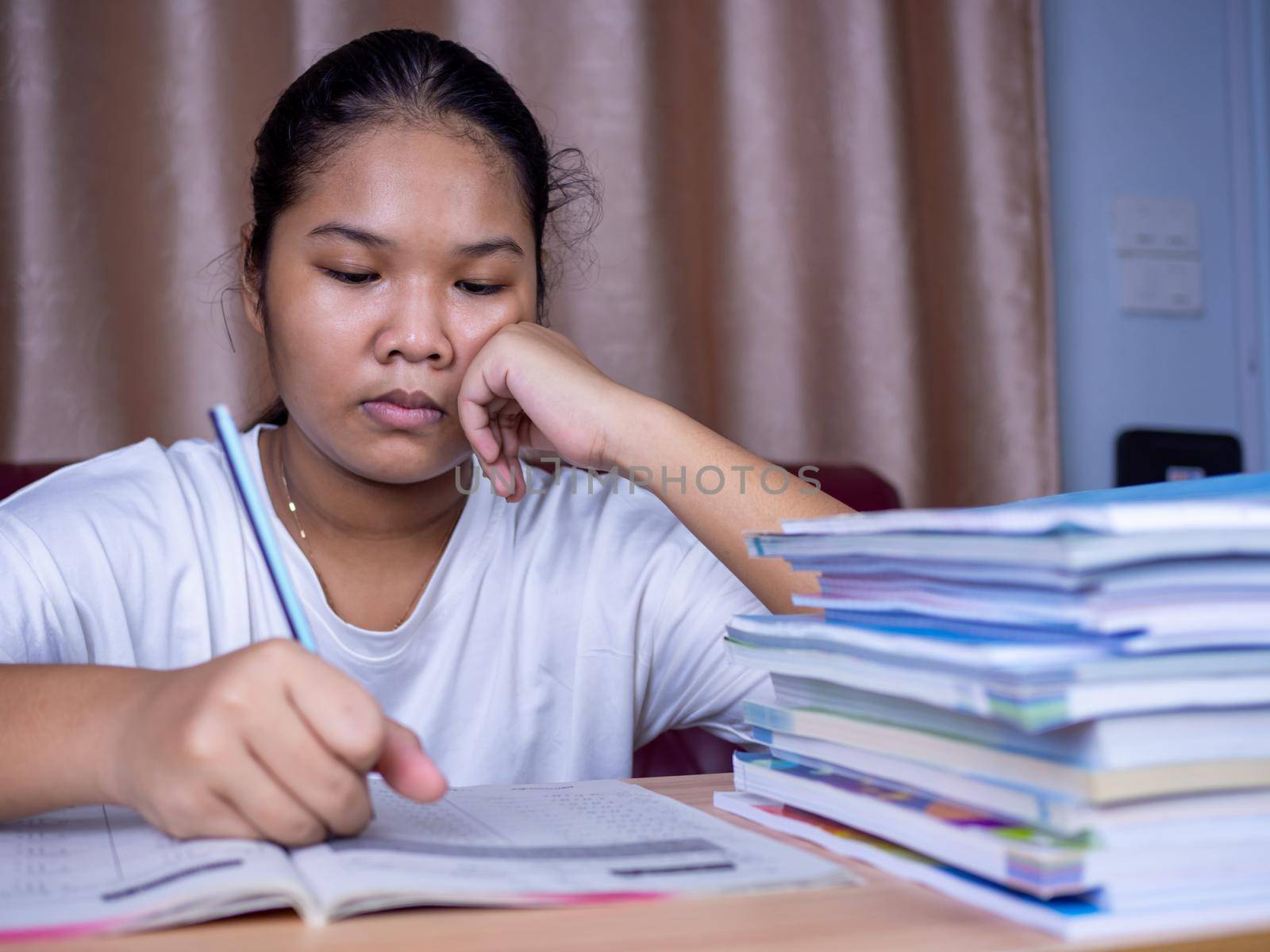 girl doing homework on a wooden table and there was a pile of books next to it The background is a red sofa and cream curtains. by Unimages2527