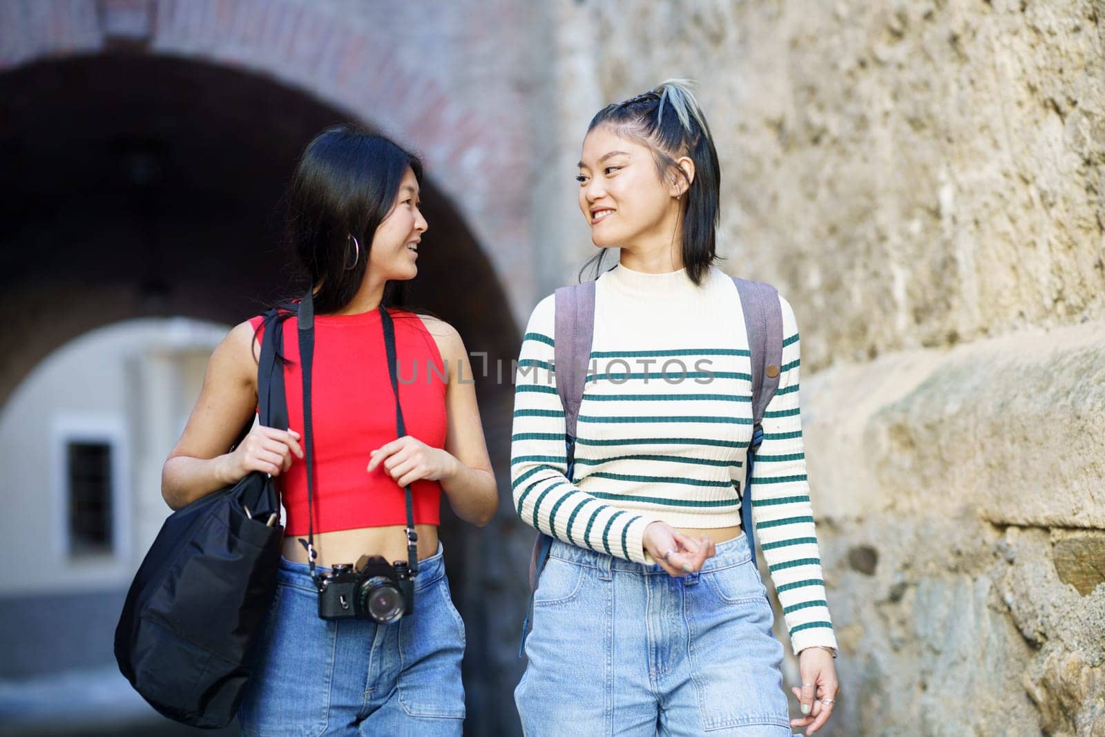 Happy young Asian female tourists in casual clothes with bags and photo camera looking at each other while strolling together near rough wall