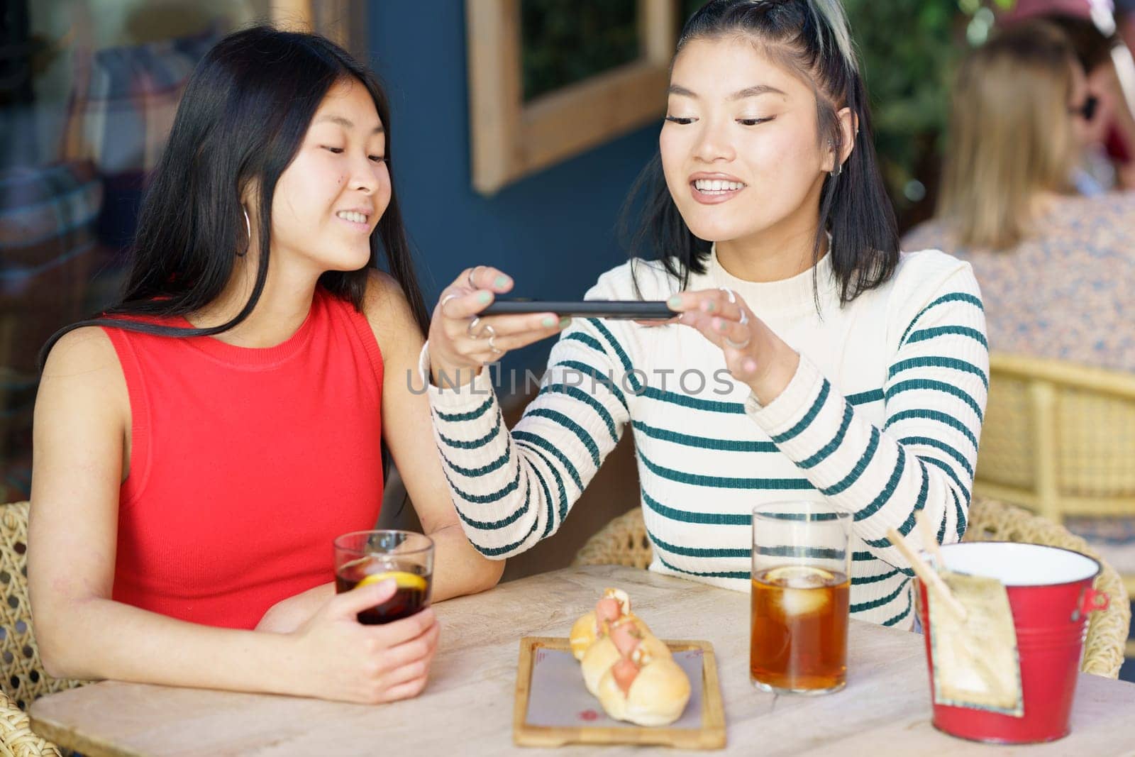 Smiling young Asian female friends, in casual clothes sitting at table with cold drinks and taking picture of tasty food on smartphone while spending time together in cafe