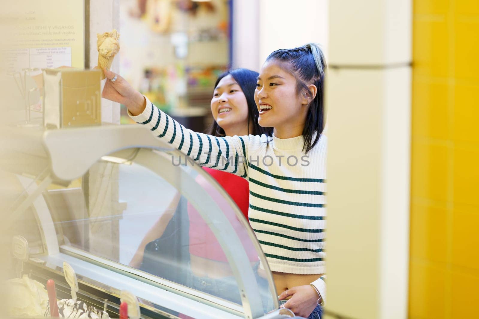 Positive young Asian girlfriends smiling happily while purchasing together ice cream in shop