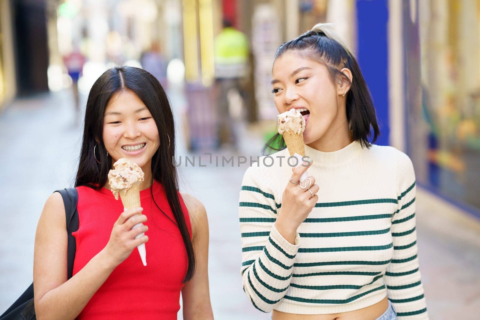 Happy Asian female friends eating delicious ice cream cone while walking together and enjoying time in city street