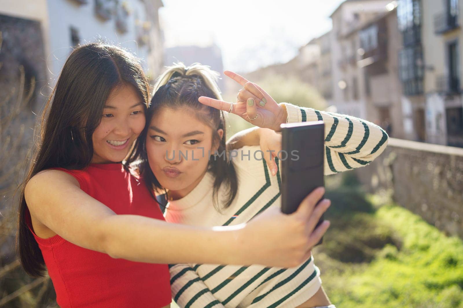 Happy Asian women in casual clothes showing rock gesture and using smartphone while shooting selfie together in Granada
