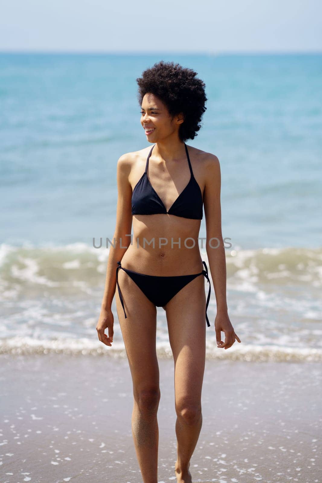 Optimistic African American female tourist in bikini looking into distance while waking on sandy coast near waving sea in tropical resort on sunny day