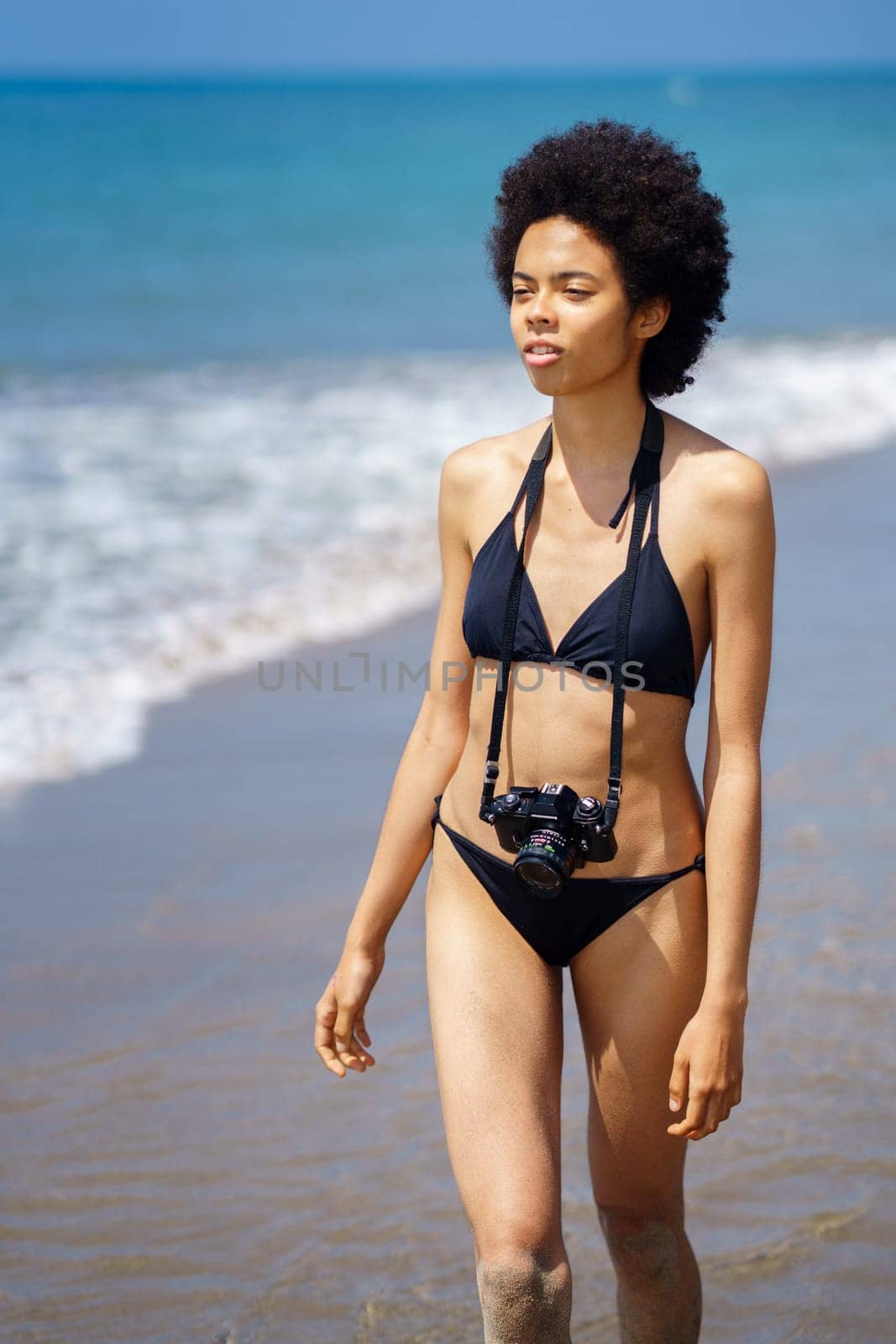 Serious African American female photographer with Afro hairstyle in swimwear with photo camera walking on shore near sea on summer day