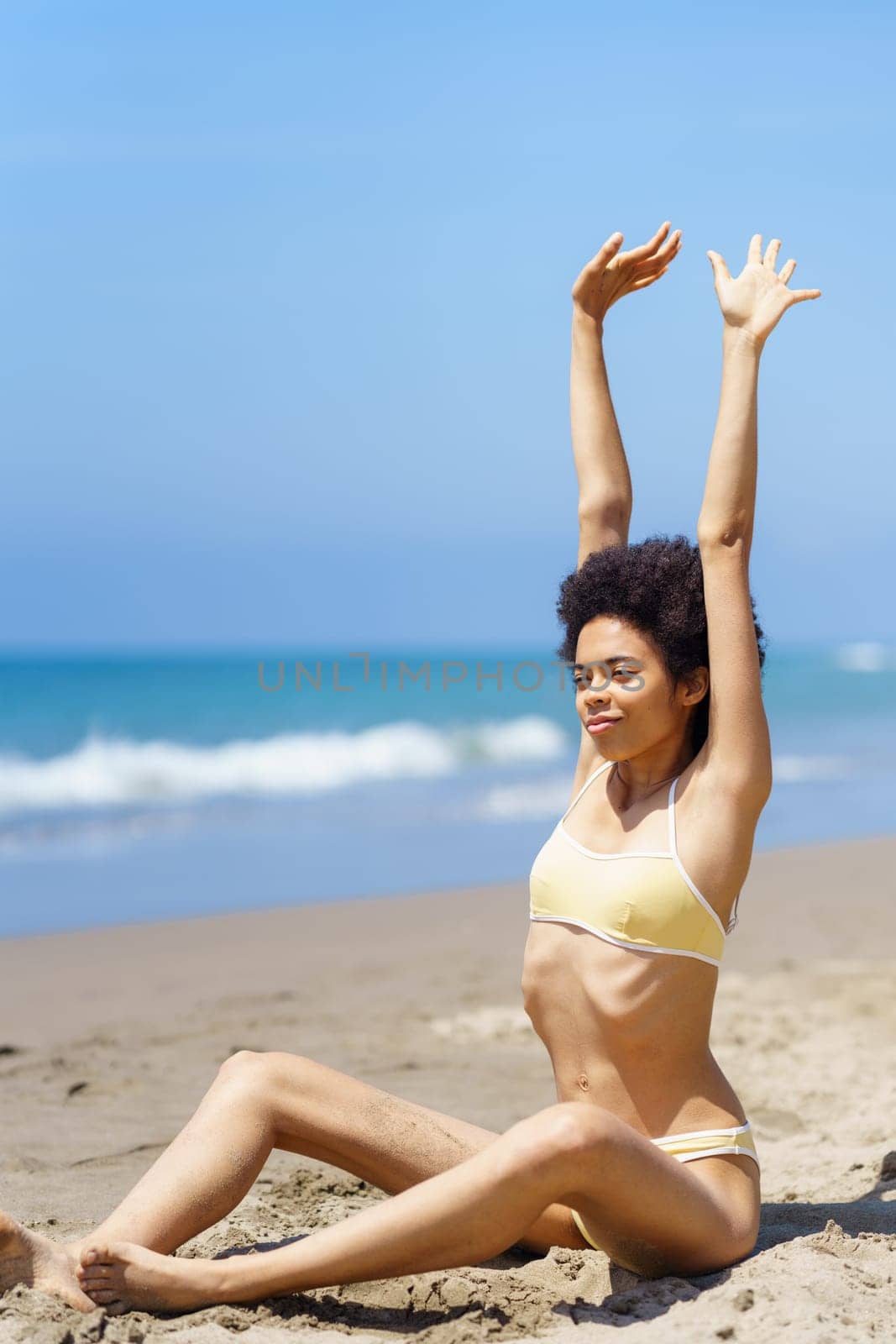 Content young fit African American female tourist with curly dark hair in stylish bikini stretching arms, while sitting on sandy beach against cloudless blue sky during summer vacation