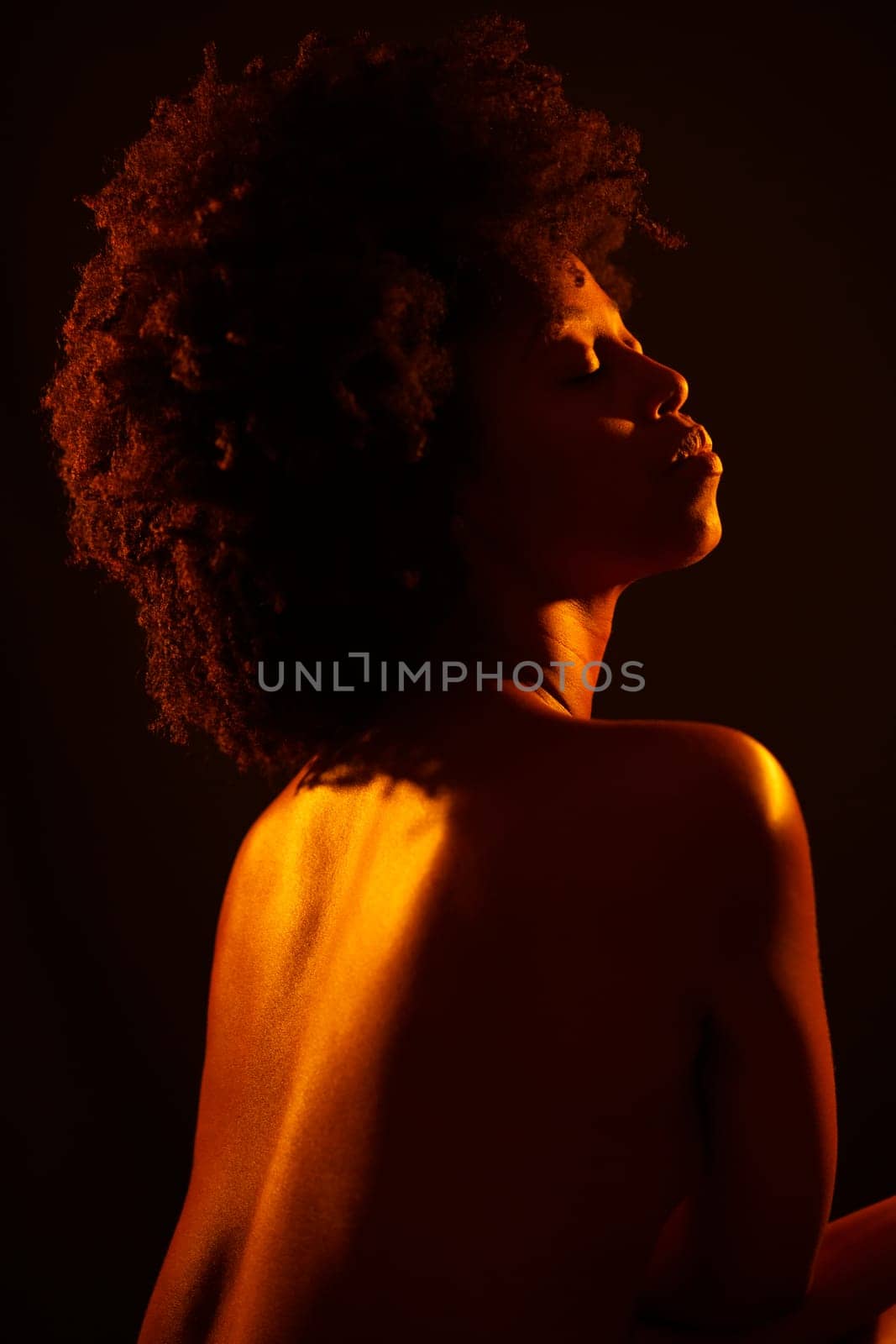 Naked black female model with Afro hairstyle closing eyes and enjoying bright neon orange light against black background
