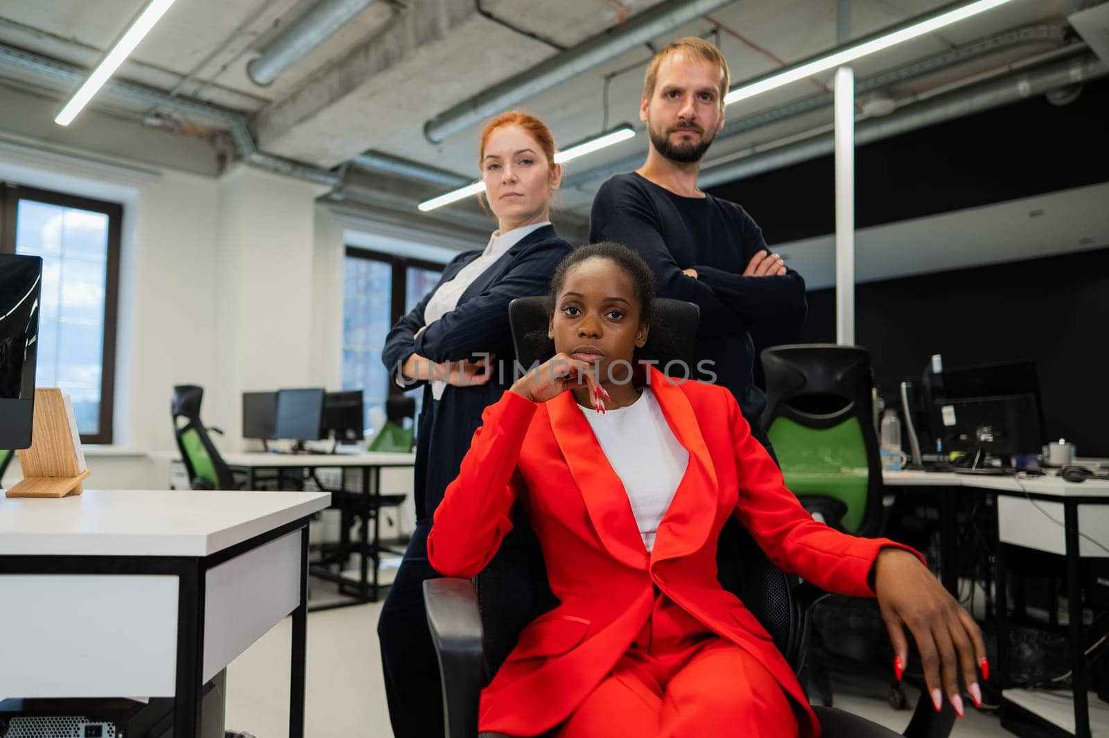 Caucasian red-haired woman, bearded Caucasian man stand behind a seated African American young woman in the office