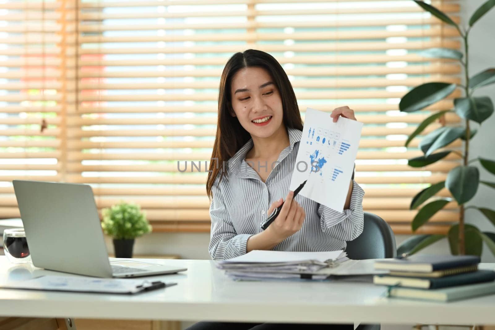 Smiling young businesswoman holding financial graph reports making virtual online presentation via laptop computer by prathanchorruangsak