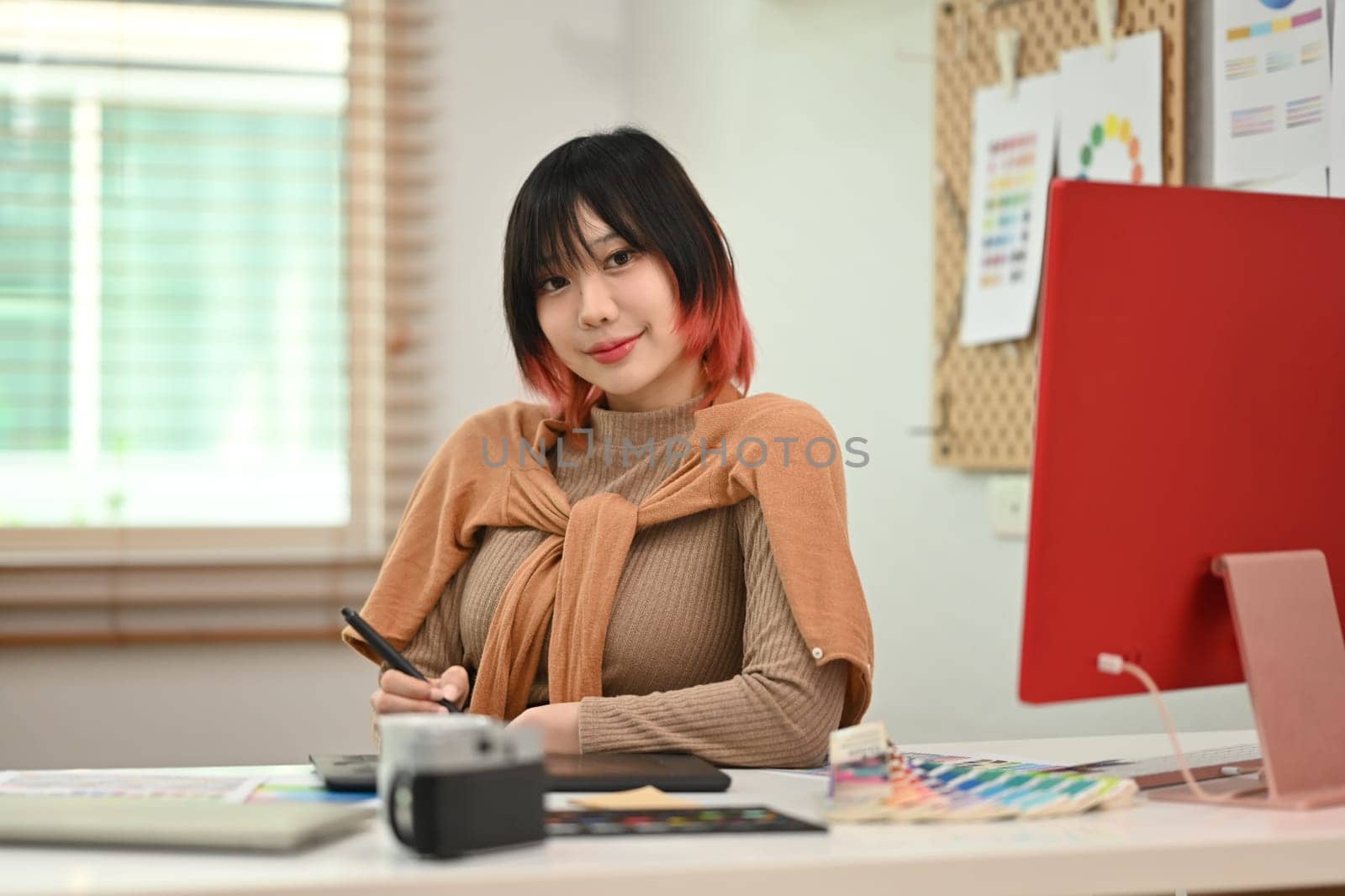 Young creative woman using graphic tablet and working with color swatch samples at modern workplace by prathanchorruangsak