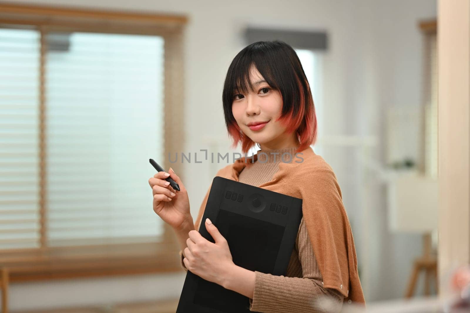 Image of stylish young woman holding graphic tablet standing in modern creative office, smiling to camera.
