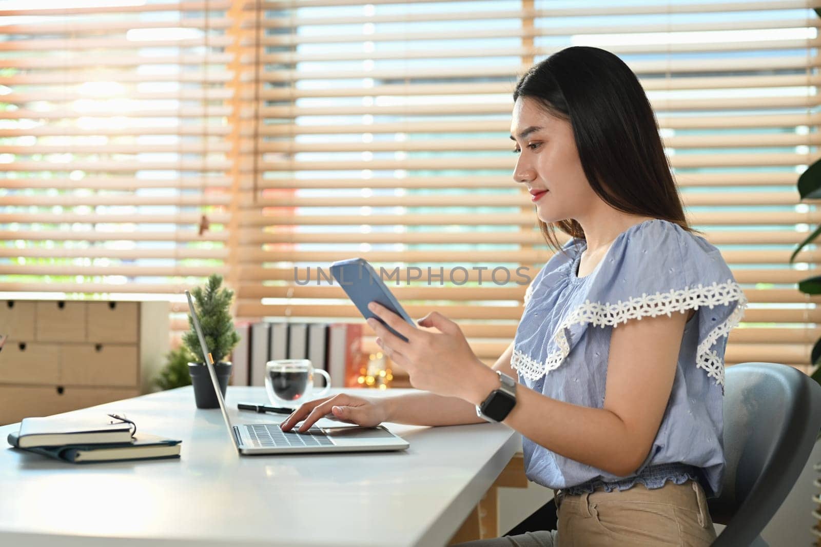 Attractive asian woman freelancer holding smart phone and using laptop at modern home office by prathanchorruangsak