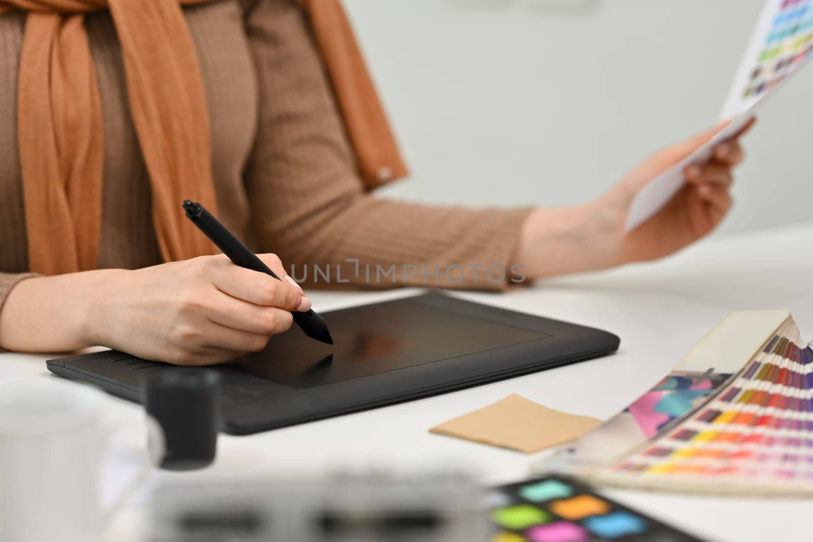 Young creative woman using graphic tablet and working with color swatch samples at modern workplace by prathanchorruangsak