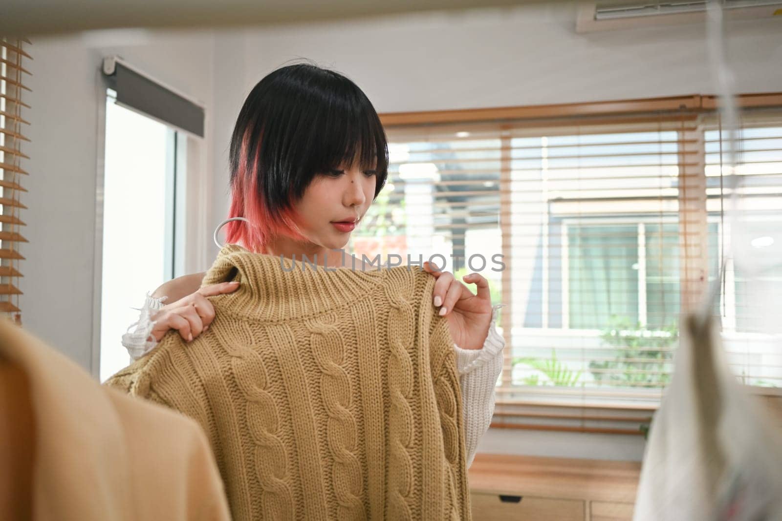 Young female fashion designer standing near clothing rack, working on new womenswear collection in modern studio by prathanchorruangsak