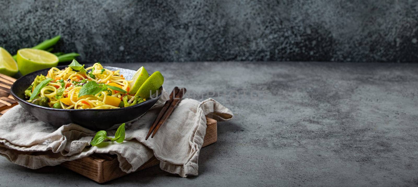 Asian vegetarian noodles with vegetables and lime in black rustic ceramic bowl, wooden chopsticks on cutting board angle view on stone background. Cooking noodles by its_al_dente