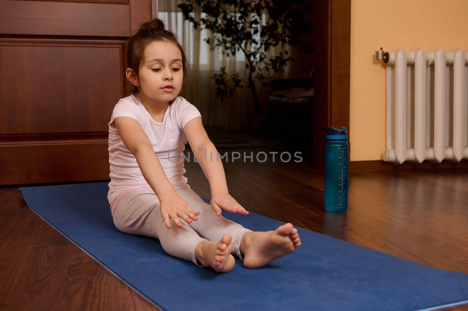Determined active little kid girl 5-6 years old, exercising barefoot on yoga mat, warming up, stretching her body, doing gymnastic exercises in cozy wooden homely atmosphere. Sport and people concept