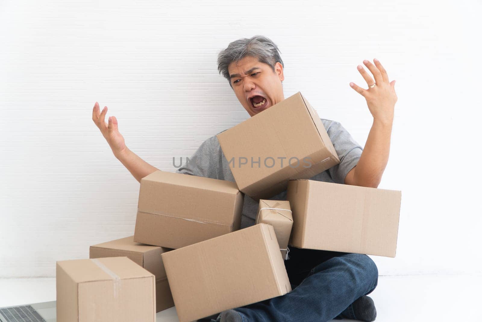 Asian Shopaholic man Shocked and sitting on the floor in the living room and Cardboard Box on top of him after The courier delivered to the home. Concept of shopping online and Shopaholic. by PattyPhoto