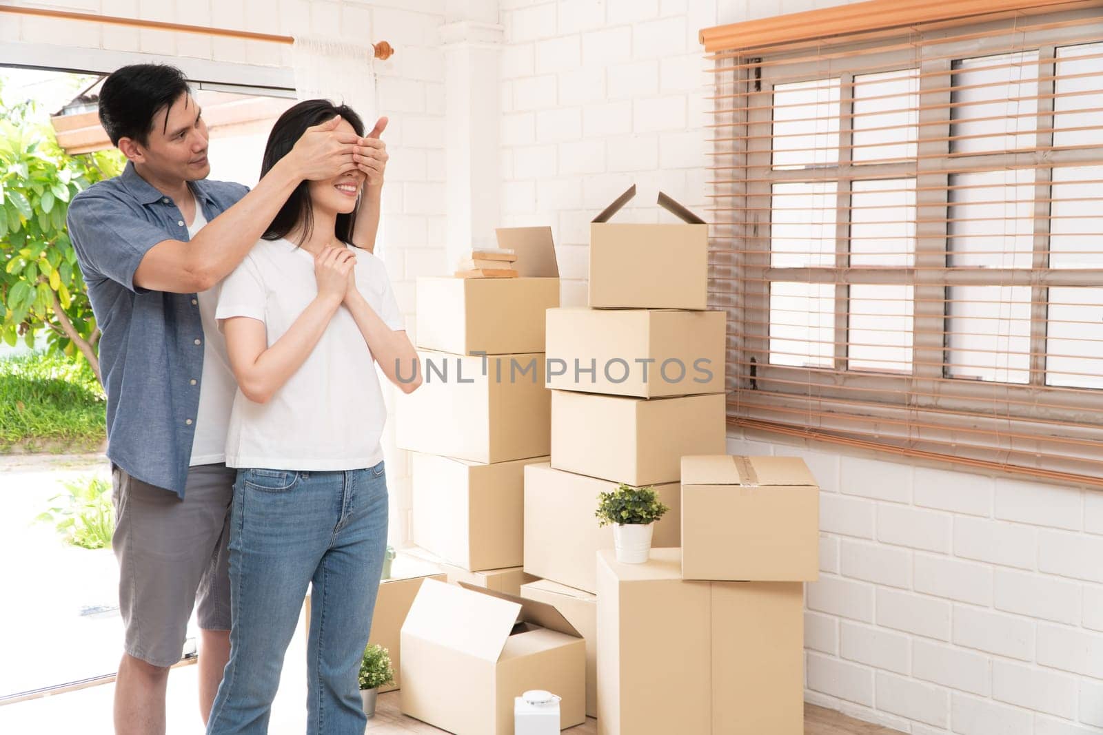 Smiling young Asian happy couple close girlfriend eyes for a surprise at moving day in their new home after buying real estate. Concept of starting a new life for a newly married couple.