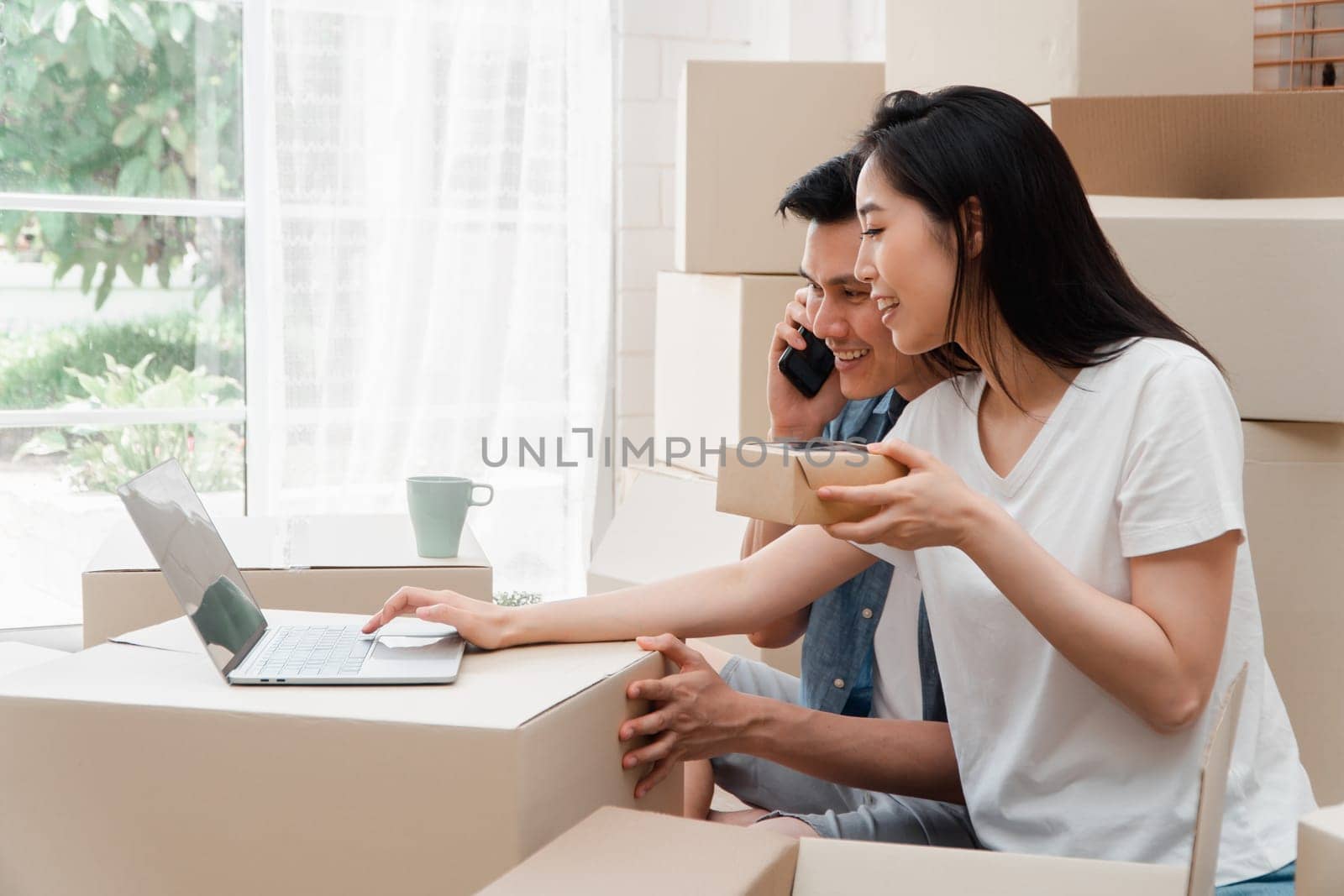 Smiling young Asian happy couple use laptop for home decoration ideas at moving day in their new home after buying real estate. Concept of starting a new life for a newly married couple.