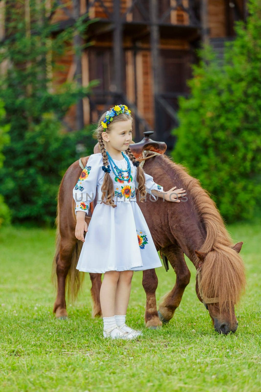 young girl in Ukrainian national dress strokes a pony that grazes on the lawn