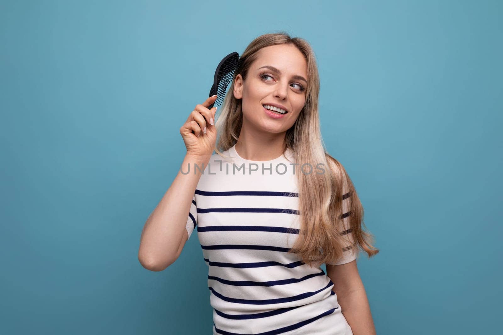 cute well-groomed european blond woman combing her hair on a blue isolated background.