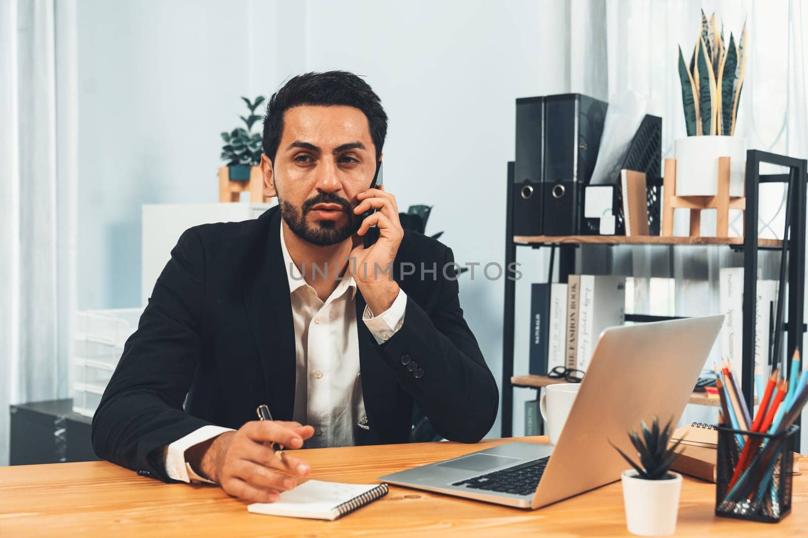 Diligent businessman busy talking on the phone call with clients while working with laptop in his office desk as concept of modern hardworking office worker lifestyle with mobile phone. Fervent