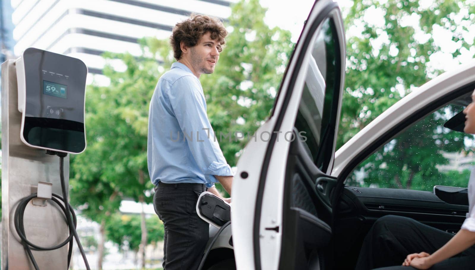 Progressive businessman and businesswoman with electric car parking and connected to public charging station before driving around city center. Eco friendly rechargeable car powered by clean energy.