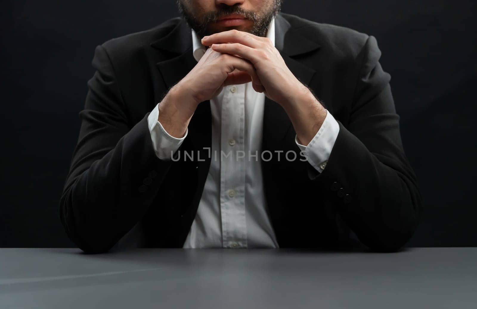 Businessman or lawyer wearing formal black suit sitting at table on isolated black background. Concept of a man with authority and seriousness gesture. equility