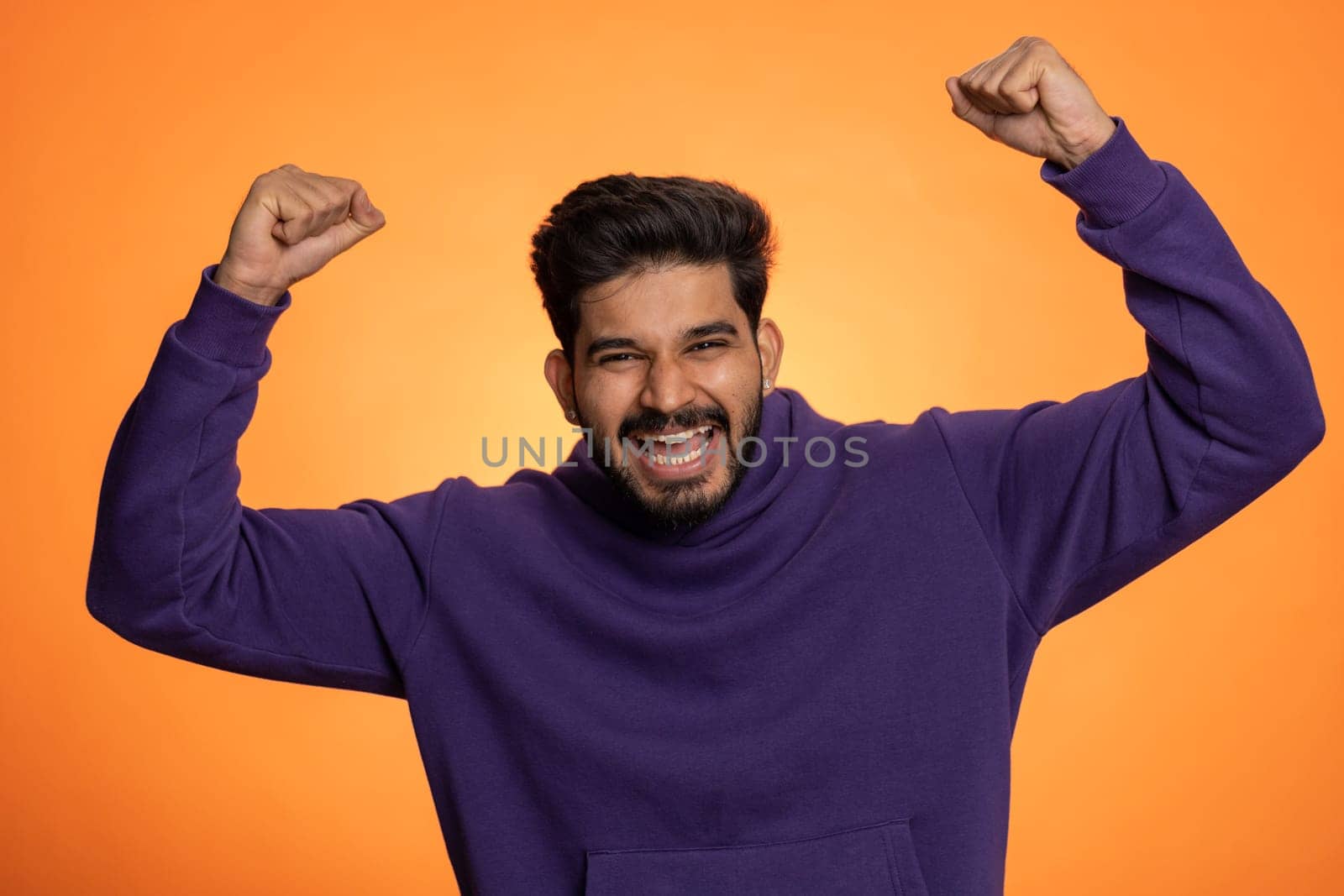 Happy joyful indian man shouting, celebrating success winning, birthday, lottery jackpot goal achievemen, good news, victory, watching TV championship. Young hindu guy on orange studio wall background