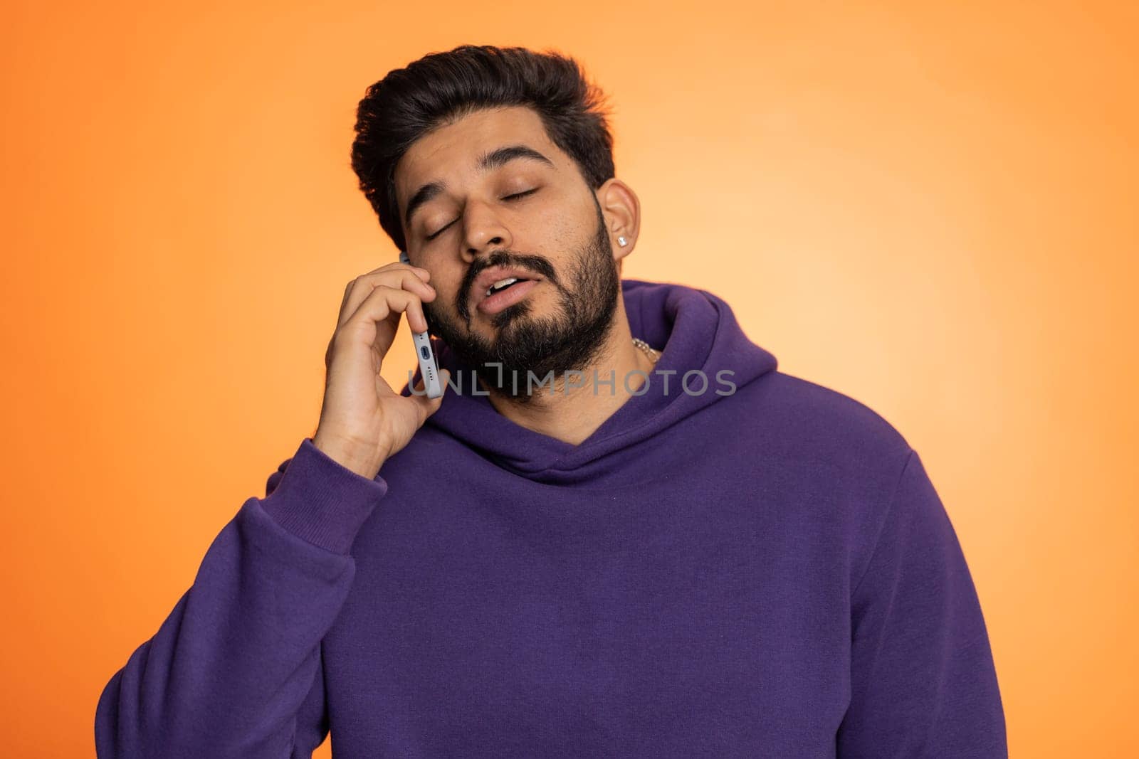 Young tired sleepy indian freelancer man talking on mobile phone with friend, making online conversation. Disinterested hindu guy having annoyed boring talk on smartphone isolated on orange background