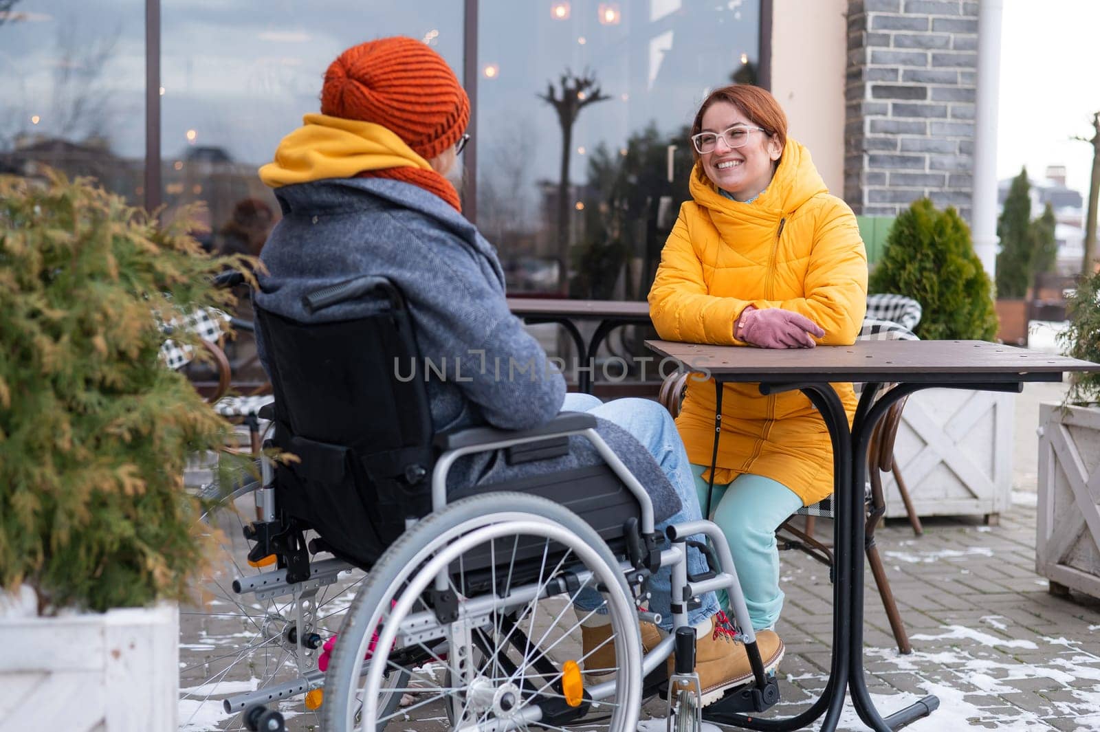 Two girlfriends in a cafe on a street terrace in winter. Woman in a wheelchair. by mrwed54